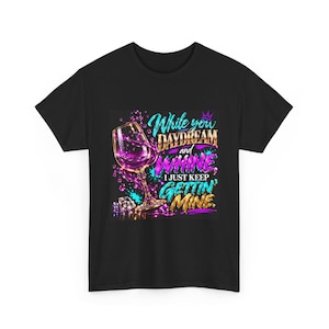 Può includere: Una t-shirt nera con la grafica di un calice da vino con liquido viola che schizza fuori, circondato da forme di diamanti. Il testo recita: While you DAYDREAM and WHINE, I JUST KEEP GETTIN' MINE. Il design utilizza colori blu, viola e oro.