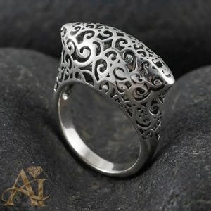Könnte beinhalten: Ein silberner Ring mit einem komplizierten, durchbrochenen Filigranmuster auf dem breiten, geschwungenen Oberteil. Das Metall hat eine polierte Oberfläche und das Design besteht aus miteinander verbundenen, wirbelnden Linien und Formen.