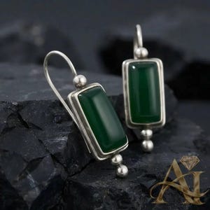 May include: A pair of silver drop earrings featuring rectangular green stones set in polished metal frames. Each earring has a curved hook wire and a small spherical bead accent at the base of the setting, creating a balanced design.