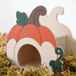 May include: A wooden pumpkin-shaped hideout for small animals, featuring a natural light wood finish, a painted orange pumpkin body, a dark brown stem, and dark green leaf accents. The structure includes a circular entrance opening.