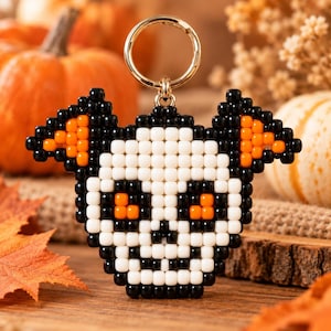 Puede incluir: Un llavero de cuentas con forma de calavera y orejas de murciélago. La calavera está hecha de cuentas blancas, negras y naranjas, con contornos negros, detalles faciales blancos y ojos y orejas de color naranja brillante, unido a una argolla dorada.