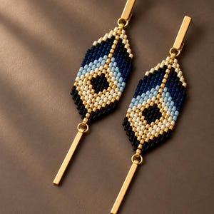 May include: A pair of drop earrings featuring a central leaf-shaped motif crafted from small beads in navy blue, light blue, cream, and black. Each earring is suspended from a gold-toned metal bar and finished with a matching gold bar at the base.
