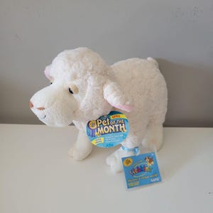 Puede incluir: Un peluche de cordero color crema con un cuerpo de tela texturizada y rizada. Una etiqueta redonda azul en el cuello dice "APRIL PET OF THE MONTH" y una etiqueta rectangular de plástico muestra el logotipo "Webkinz" y el texto "discover a virtual world".