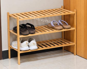 May include: A three-tier bamboo shoe rack with a slatted design and the NestivoHome logo printed on the frame. The rack holds a pair of black sandals, a pair of light purple flip-flops, and a pair of white athletic sneakers on its shelves.