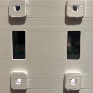 Puede incluir: Una placa de pared blanca impresa en 3D con dos aberturas rectangulares y cuatro puntos de montaje cuadrados, cada uno con un orificio central. La parte superior muestra un logotipo y la inferior el texto en relieve Made in the USA.
