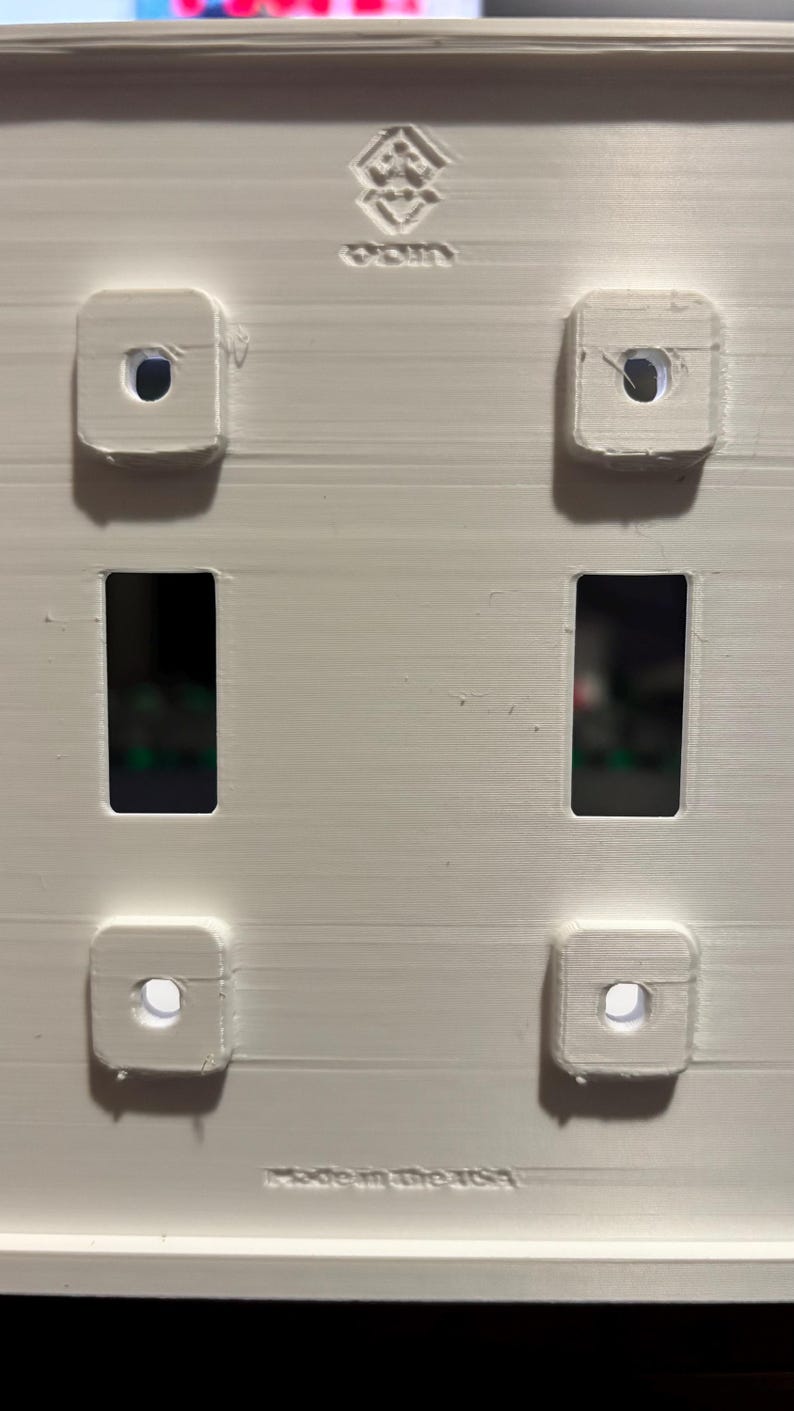 Puede incluir: Una placa de pared blanca impresa en 3D con dos aberturas rectangulares y cuatro puntos de montaje cuadrados, cada uno con un orificio central. La parte superior muestra un logotipo y la inferior el texto en relieve Made in the USA.