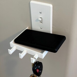Puede incluir: Un estante de pared de pl&aacute;stico blanco dise&ntilde;ado para fijarse a una placa de interruptor de luz est&aacute;ndar. El estante cuenta con una superficie rectangular plana que sostiene un objeto negro y tres ganchos peque&ntilde;os para colgar llaves.