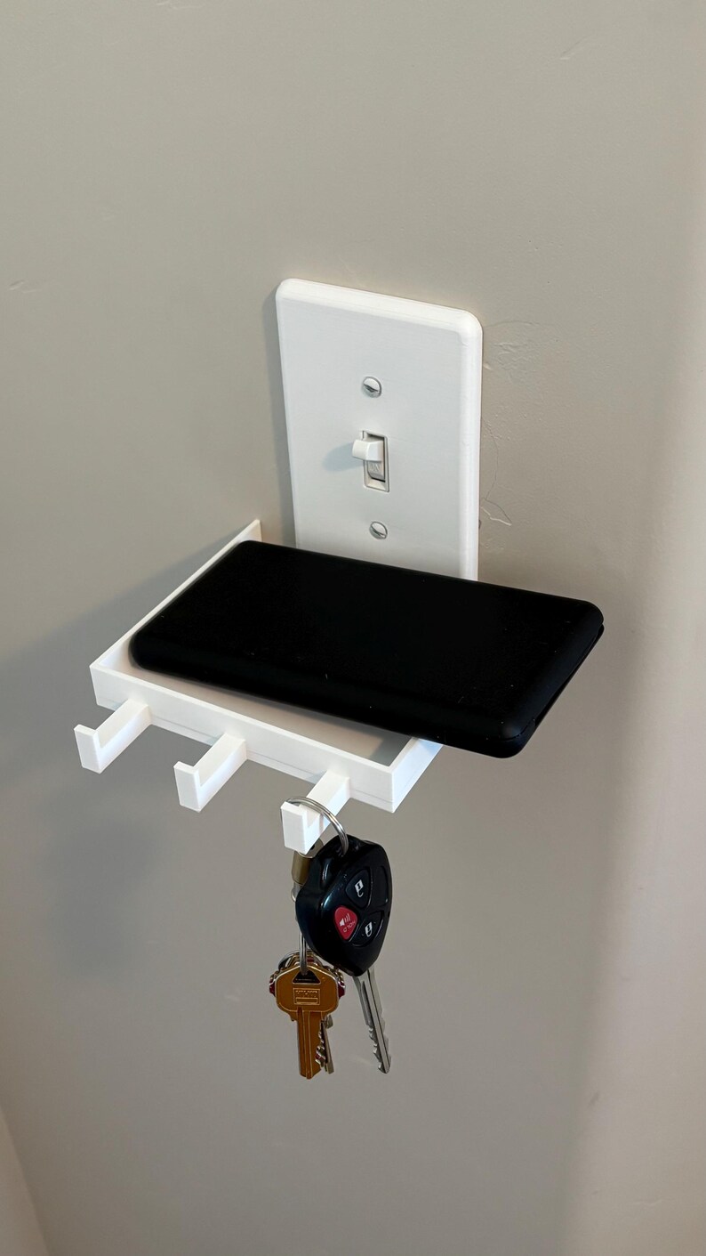 Puede incluir: Un estante de pared de pl&aacute;stico blanco dise&ntilde;ado para fijarse a una placa de interruptor de luz est&aacute;ndar. El estante cuenta con una superficie rectangular plana que sostiene un objeto negro y tres ganchos peque&ntilde;os para colgar llaves.
