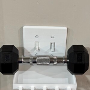 Puede incluir: Un estante de pared de pl&aacute;stico blanco dise&ntilde;ado para encajar sobre una placa de interruptor doble. El estante cuenta con una superficie rectangular plana y cinco ganchos peque&ntilde;os en el borde inferior, sosteniendo una mancuerna negra y plateada.