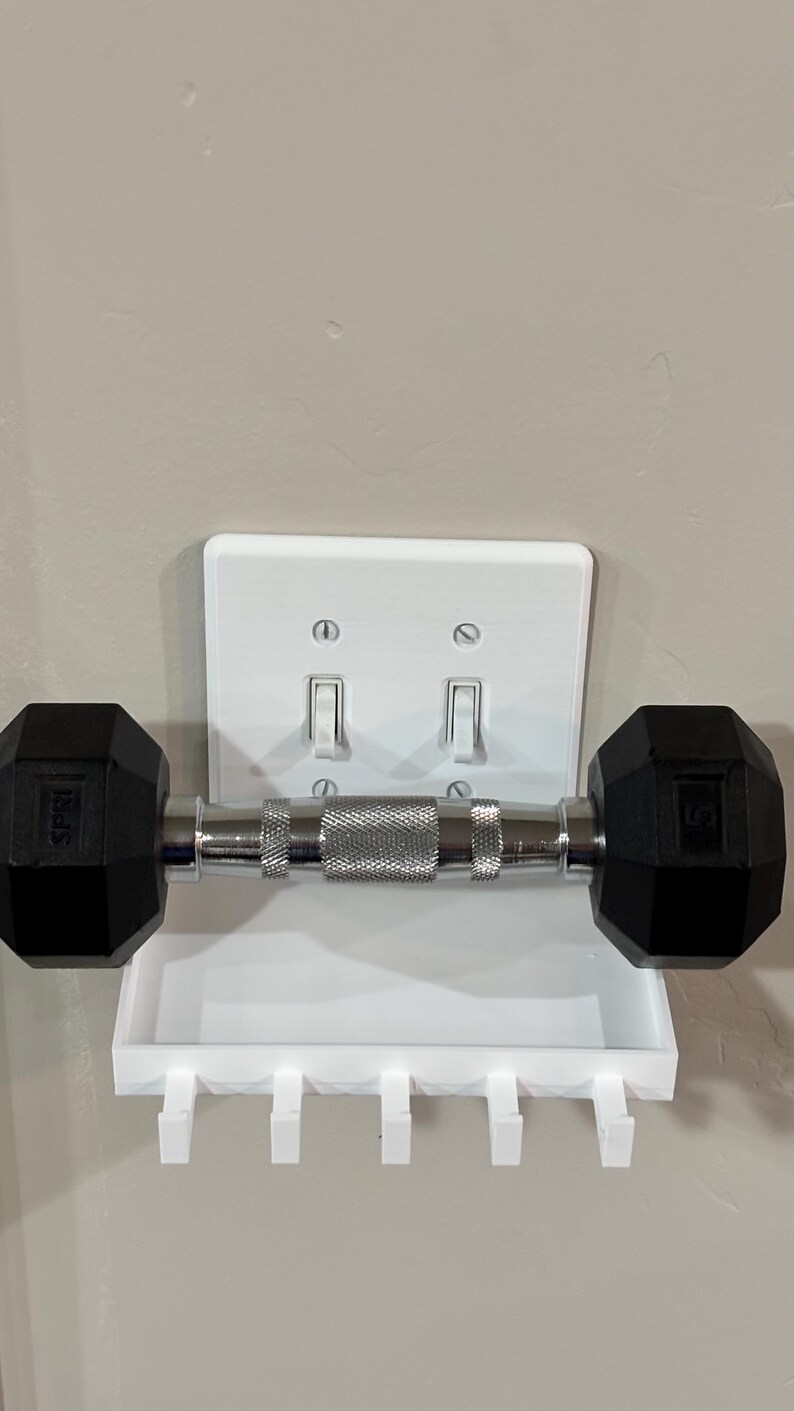 Puede incluir: Un estante de pared de pl&aacute;stico blanco dise&ntilde;ado para encajar sobre una placa de interruptor doble. El estante cuenta con una superficie rectangular plana y cinco ganchos peque&ntilde;os en el borde inferior, sosteniendo una mancuerna negra y plateada.