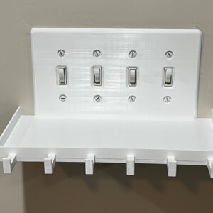 Puede incluir: Una placa de pared blanca para cuatro interruptores de luz, con un estante horizontal integrado y seis peque&ntilde;os ganchos en el borde inferior. La placa rectangular est&aacute; asegurada con ocho tornillos visibles y aloja cuatro interruptores.
