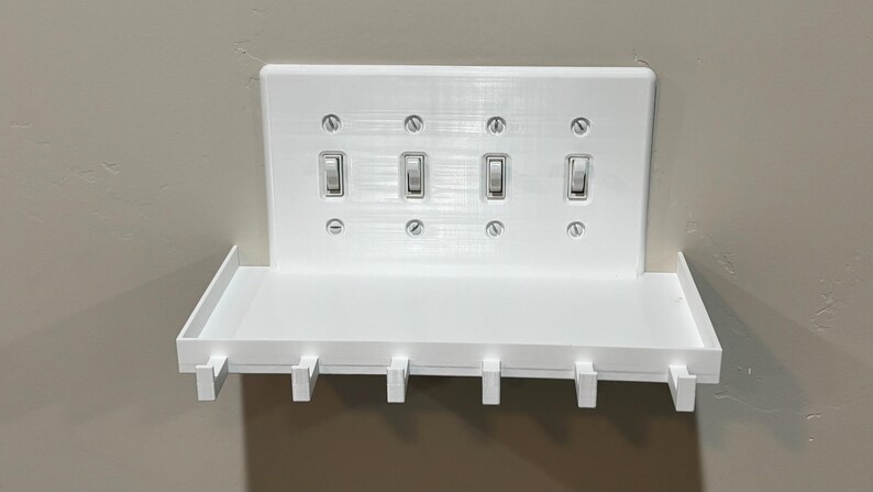 Puede incluir: Una placa de pared blanca para cuatro interruptores de luz, con un estante horizontal integrado y seis peque&ntilde;os ganchos en el borde inferior. La placa rectangular est&aacute; asegurada con ocho tornillos visibles y aloja cuatro interruptores.