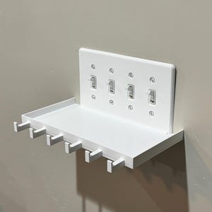 Puede incluir: Un estante de pared blanco con una placa de cubierta integrada para cuatro interruptores de luz. El estante incluye una superficie rectangular plana y cinco peque&ntilde;os ganchos que se extienden desde el borde frontal para organizar objetos.