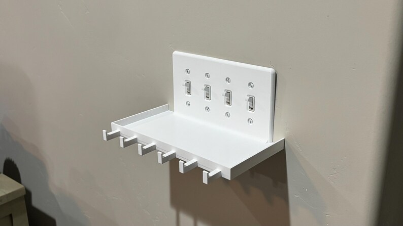Puede incluir: Un estante de pared blanco con una placa de cubierta integrada para cuatro interruptores de luz. El estante incluye una superficie rectangular plana y cinco peque&ntilde;os ganchos que se extienden desde el borde frontal para organizar objetos.