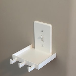 Puede incluir: Un estante de pared de pl&aacute;stico blanco dise&ntilde;ado para encajar sobre un interruptor de luz est&aacute;ndar. El estante rectangular cuenta con una superficie plana y tres peque&ntilde;os ganchos en el borde frontal para colgar objetos.