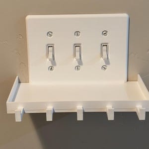 Puede incluir: Una placa de cubierta blanca para triple interruptor de luz con un estante horizontal integrado y cinco ganchos orientados hacia abajo. La placa rectangular est&aacute; asegurada con seis tornillos visibles y aloja tres interruptores de palanca.