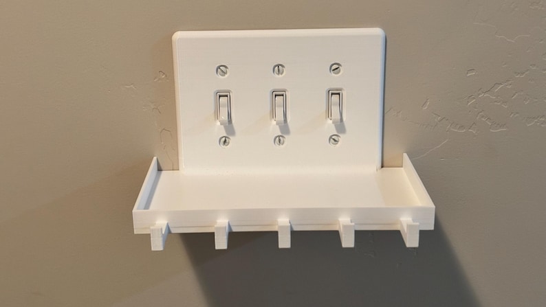 Puede incluir: Una placa de cubierta blanca para triple interruptor de luz con un estante horizontal integrado y cinco ganchos orientados hacia abajo. La placa rectangular est&aacute; asegurada con seis tornillos visibles y aloja tres interruptores de palanca.