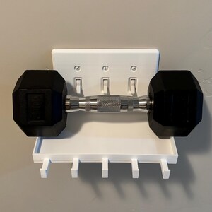 Puede incluir: Un estante de pared blanco dise&ntilde;ado para encajar sobre una placa de interruptor triple, con una superficie superior plana y cinco ganchos peque&ntilde;os debajo. Una mancuerna hexagonal negra y plateada descansa horizontalmente sobre el estante.