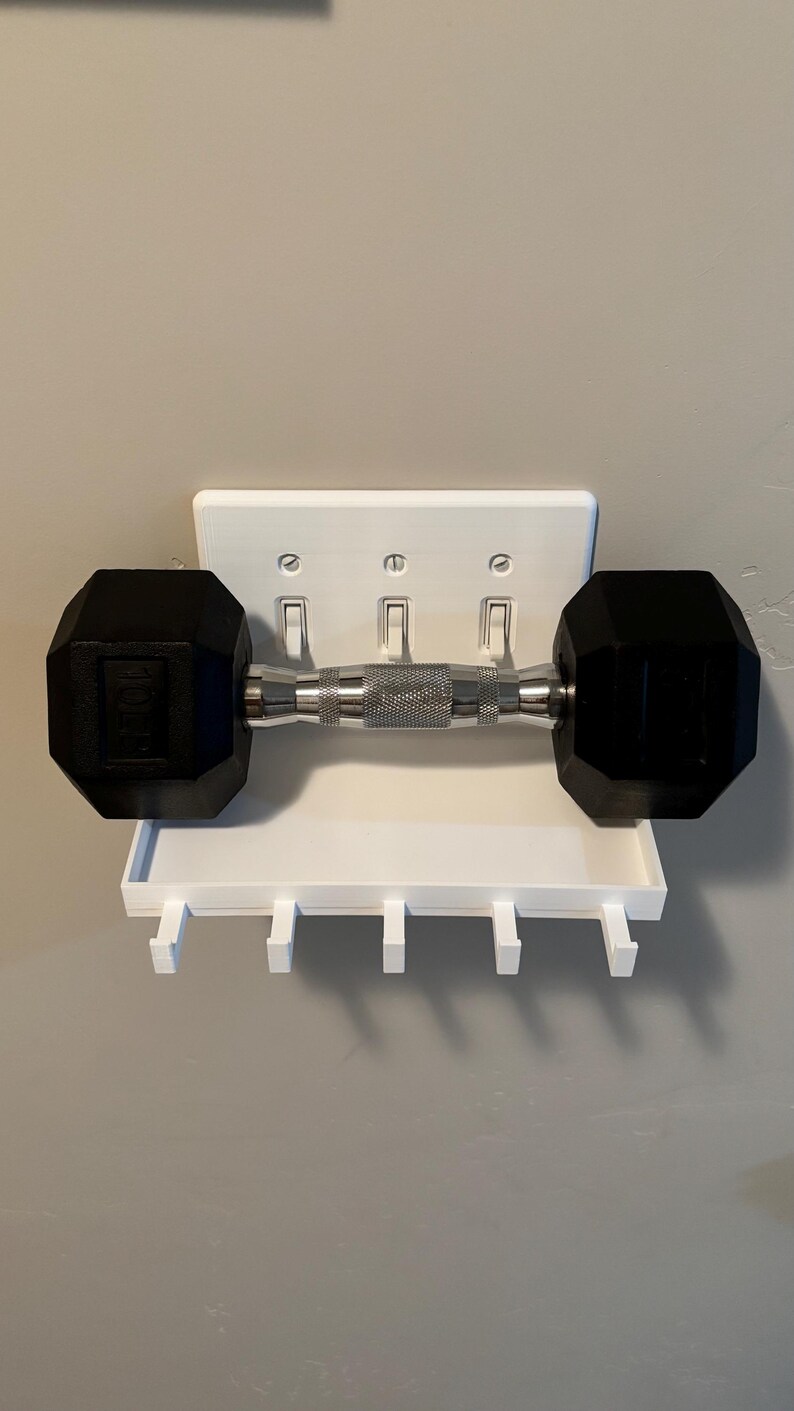 Puede incluir: Un estante de pared blanco dise&ntilde;ado para encajar sobre una placa de interruptor triple, con una superficie superior plana y cinco ganchos peque&ntilde;os debajo. Una mancuerna hexagonal negra y plateada descansa horizontalmente sobre el estante.