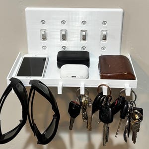 Puede incluir: Un estante organizador de pared blanco con una placa de cubierta para interruptor de luz cu&aacute;druple. El estante sostiene un tel&eacute;fono inteligente, dos estuches rectangulares peque&ntilde;os y una billetera, con ganchos inferiores para gafas y llaves.