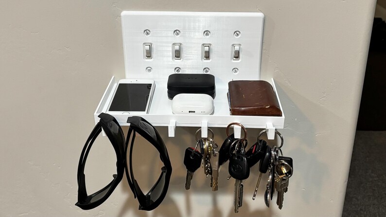 Puede incluir: Un estante organizador de pared blanco con una placa de cubierta para interruptor de luz cu&aacute;druple. El estante sostiene un tel&eacute;fono inteligente, dos estuches rectangulares peque&ntilde;os y una billetera, con ganchos inferiores para gafas y llaves.