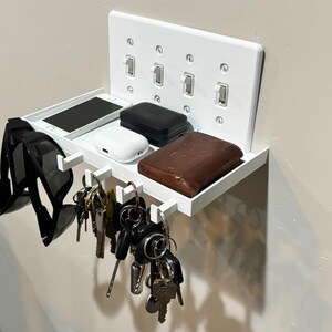 Puede incluir: Un organizador de pared blanco con un estante rectangular y cuatro ganchos integrados. La placa trasera se asemeja a una cubierta de interruptor de luz cu&aacute;druple. El estante sostiene un tel&eacute;fono inteligente, un estuche blanco, uno negro y una cartera de cuero marr&oacute;n.