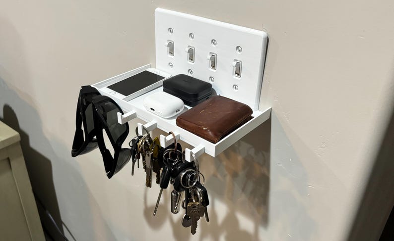 Puede incluir: Un organizador de pared blanco con un estante rectangular y cuatro ganchos integrados. La placa trasera se asemeja a una cubierta de interruptor de luz cu&aacute;druple. El estante sostiene un tel&eacute;fono inteligente, un estuche blanco, uno negro y una cartera de cuero marr&oacute;n.