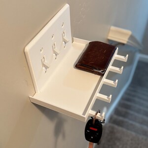 Puede incluir: Un estante de pared blanco dise&ntilde;ado para encajar sobre una placa de interruptor triple. El estante cuenta con una superficie plana que sostiene una cartera de cuero marr&oacute;n e incluye ganchos integrados en la parte inferior para colgar llaves.