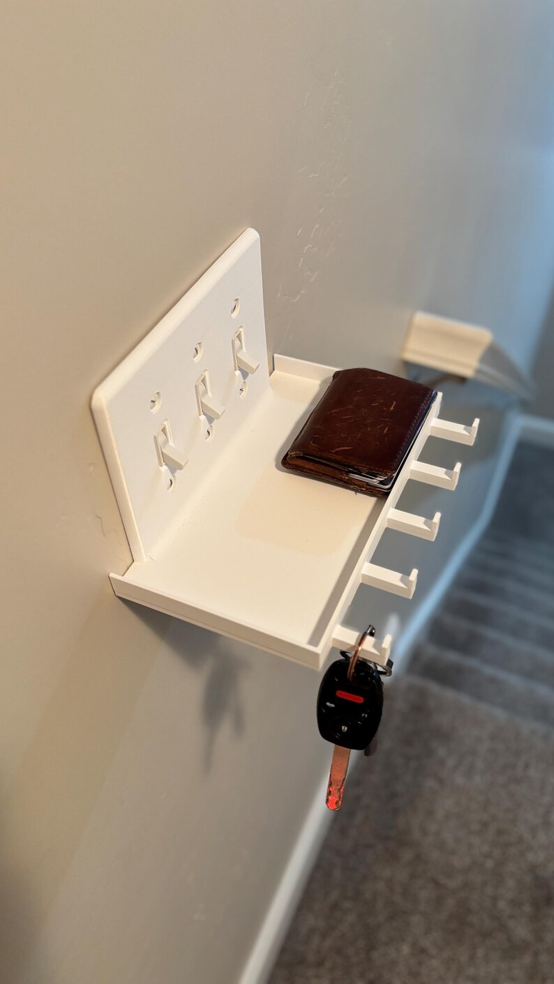 Puede incluir: Un estante de pared blanco dise&ntilde;ado para encajar sobre una placa de interruptor triple. El estante cuenta con una superficie plana que sostiene una cartera de cuero marr&oacute;n e incluye ganchos integrados en la parte inferior para colgar llaves.