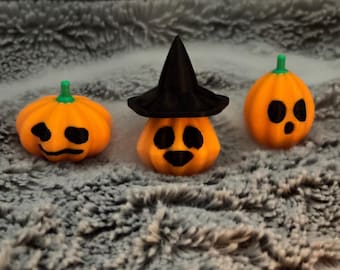 Puede incluir: Tres pequeñas figuras de calabaza de color naranja con tallos verdes y rasgos faciales pintados en negro. La calabaza central lleva un sombrero de bruja negro puntiagudo. Cada calabaza tiene una textura acanalada y una forma redondeada.