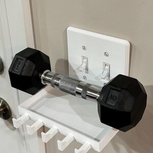 Puede incluir: Un estante organizador de pared de pl&aacute;stico blanco con cuatro ganchos, colocado directamente sobre una placa de interruptor doble. Una mancuerna de metal negro y plata con el texto SPRI y 5 impreso en los extremos descansa sobre el estante.