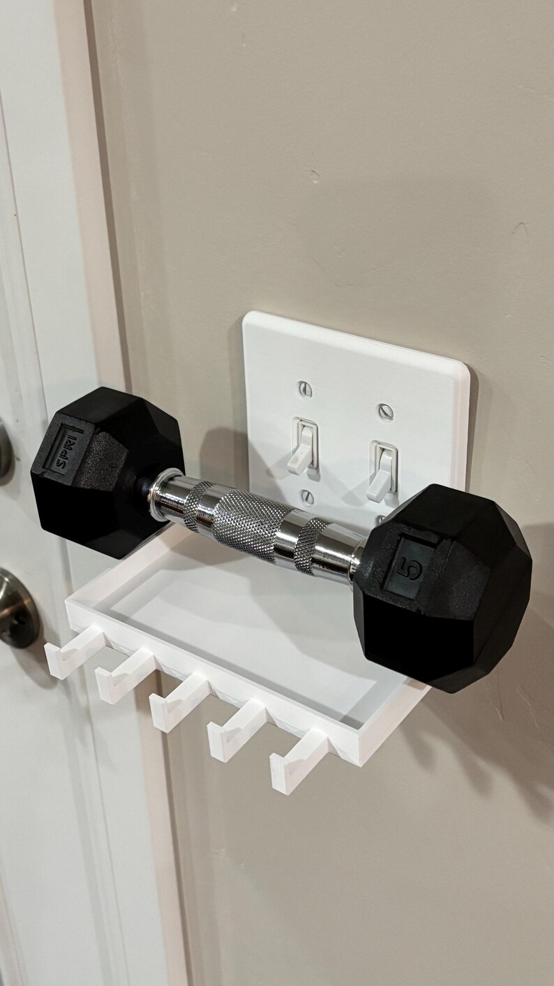 Puede incluir: Un estante organizador de pared de pl&aacute;stico blanco con cuatro ganchos, colocado directamente sobre una placa de interruptor doble. Una mancuerna de metal negro y plata con el texto SPRI y 5 impreso en los extremos descansa sobre el estante.
