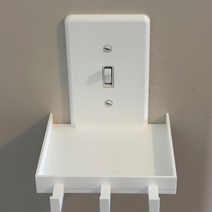 Puede incluir: Un estante de pared blanco dise&ntilde;ado para encajar sobre una placa de interruptor de luz est&aacute;ndar. El estante rectangular cuenta con una superficie plana con tres peque&ntilde;os ganchos en el borde frontal, fijado con dos tornillos a la placa.