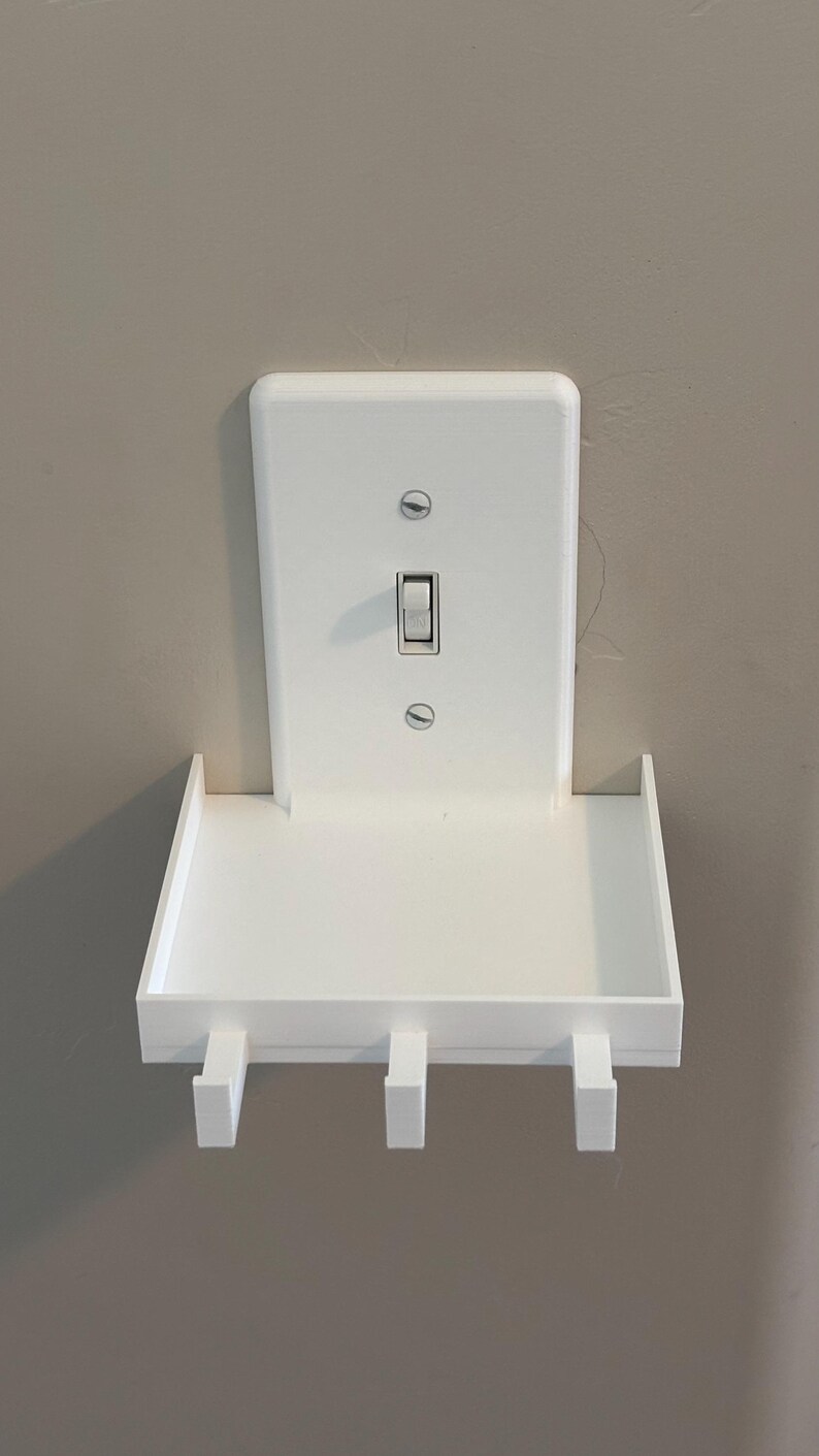 Puede incluir: Un estante de pared blanco dise&ntilde;ado para encajar sobre una placa de interruptor de luz est&aacute;ndar. El estante rectangular cuenta con una superficie plana con tres peque&ntilde;os ganchos en el borde frontal, fijado con dos tornillos a la placa.