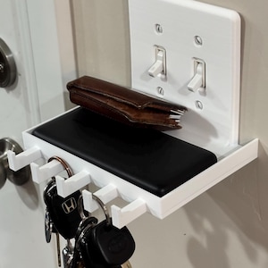 Puede incluir: Un estante de pared de pl&aacute;stico blanco integrado con una placa de interruptor doble. El estante cuenta con una superficie plana que sostiene una cartera de cuero marr&oacute;n y un objeto rectangular negro, con cinco ganchos debajo para llaves.