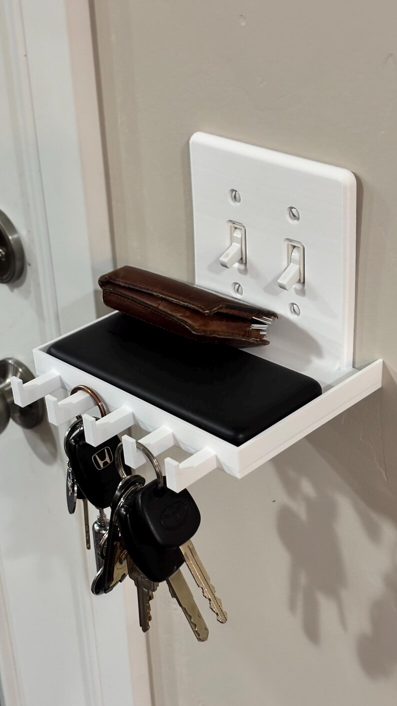 Puede incluir: Un estante de pared de pl&aacute;stico blanco integrado con una placa de interruptor doble. El estante cuenta con una superficie plana que sostiene una cartera de cuero marr&oacute;n y un objeto rectangular negro, con cinco ganchos debajo para llaves.