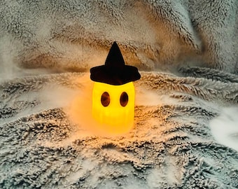 Puede incluir: Una lámpara brillante con forma de fantasma amarillo que lleva un sombrero puntiagudo negro. El fantasma tiene dos ojos ovalados oscuros y emite una luz cálida, rodeado por un ligero efecto de niebla o bruma, creando una iluminación suave.