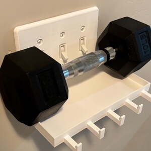 Puede incluir: Un estante de pared de pl&aacute;stico blanco con cinco ganchos, fijado sobre una placa de interruptor triple. Una mancuerna hexagonal negra de 10 lb con un mango de metal plateado texturizado descansa horizontalmente sobre el estante.