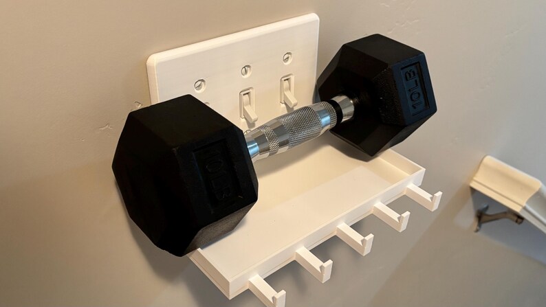 Puede incluir: Un estante de pared de pl&aacute;stico blanco con cinco ganchos, fijado sobre una placa de interruptor triple. Una mancuerna hexagonal negra de 10 lb con un mango de metal plateado texturizado descansa horizontalmente sobre el estante.