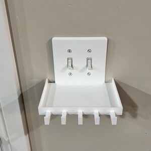 Puede incluir: Un estante de pared de pl&aacute;stico blanco dise&ntilde;ado para encajar sobre una placa de interruptor doble. El estante rectangular cuenta con una superficie plana y cinco ganchos orientados hacia abajo en el borde frontal, fijados con los tornillos del interruptor.
