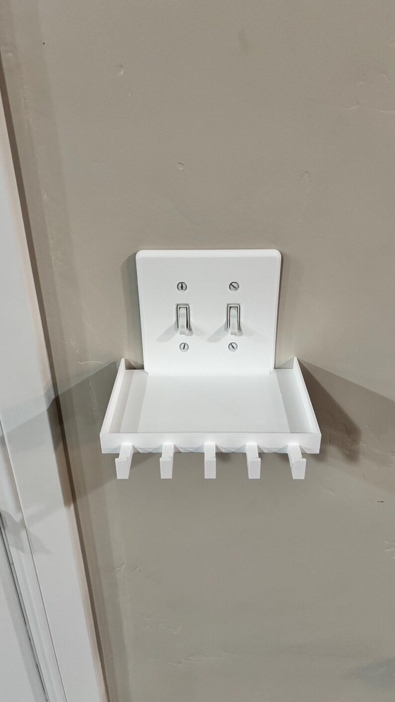 Puede incluir: Un estante de pared de pl&aacute;stico blanco dise&ntilde;ado para encajar sobre una placa de interruptor doble. El estante rectangular cuenta con una superficie plana y cinco ganchos orientados hacia abajo en el borde frontal, fijados con los tornillos del interruptor.