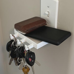 Puede incluir: Un estante de pared de pl&aacute;stico blanco fijado a una placa de interruptor de luz. El estante sostiene una cartera de cuero marr&oacute;n y un tel&eacute;fono inteligente negro. Tres peque&ntilde;os ganchos en el borde frontal sostienen llaves de metal.