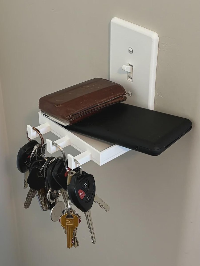 Puede incluir: Un estante de pared de pl&aacute;stico blanco fijado a una placa de interruptor de luz. El estante sostiene una cartera de cuero marr&oacute;n y un tel&eacute;fono inteligente negro. Tres peque&ntilde;os ganchos en el borde frontal sostienen llaves de metal.