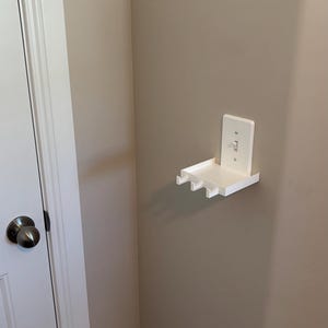 Puede incluir: Un estante de pared de pl&aacute;stico blanco dise&ntilde;ado para encajar sobre una placa de interruptor de luz est&aacute;ndar. El estante cuenta con una superficie rectangular plana con tres peque&ntilde;os ganchos que sobresalen del borde frontal.