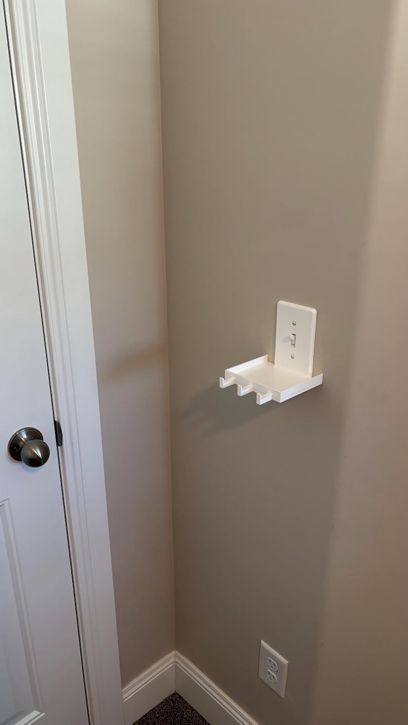 Puede incluir: Un estante de pared de pl&aacute;stico blanco dise&ntilde;ado para encajar sobre una placa de interruptor de luz est&aacute;ndar. El estante cuenta con una superficie rectangular plana con tres peque&ntilde;os ganchos que sobresalen del borde frontal.