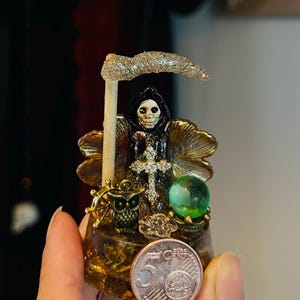 May include: A small resin sculpture featuring a hooded skeleton figure holding a glittery scythe, adorned with a crystal cross, a bronze owl, and a green sphere. A 5 Euro cent coin is placed in front for scale, showing the object's compact size.