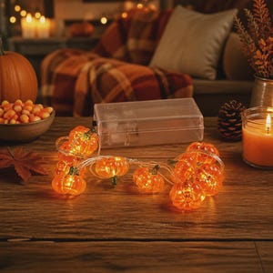 Pode incluir: Um fio de luzes LED em forma de abóbora laranja, ligado por um fio fino a uma caixa de pilhas retangular de plástico transparente. As pequenas abóboras translúcidas estão iluminadas, emitindo um brilho quente sobre a superfície.