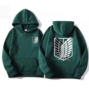 Könnte beinhalten: Ein dunkelgrüner Kapuzenpullover mit einer Kängurutasche auf der Vorderseite und langen Ärmeln. Auf der linken Brust befindet sich ein kleines weiß-schwarzes Flügel-Emblem, während der Rücken ein großes, weiß-schwarzes Flügelschild-Motiv zeigt.