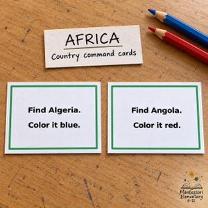 Op de afbeelding: Drie witte rechthoekige kaarten met groene randen en zwarte tekst. Eén kaart leest AFRICA Country command cards. De tweede kaart leest Find Algeria. Color it blue. De derde kaart leest Find Angola. Color it red. Een blauw en rood potlood.