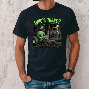 May include: A black short-sleeved t-shirt featuring a graphic print of a green alien in bed under a blanket with the text "WHO'S THERE?" above it. The scene shows an astronaut looking through a window at the alien, with a digital clock reading 3:03.